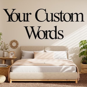 May include: A bedroom scene featuring a bed with neutral-toned bedding and a wooden frame. The wall above the bed displays the text "Your Custom Words" in large, black lettering. Decorative plants and a small table with a clock add to the room's aesthetic.