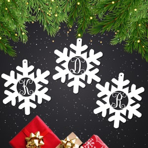 May include: Three white snowflake ornaments with a circle in the center for a monogram. The snowflakes are on a black background. The monograms are K, D, and R.