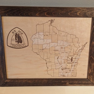 May include: Wooden framed map of Wisconsin with the Ice Age National Scenic Trail highlighted. The map is laser-cut into a light-colored wood, with a dark brown stained frame. The Ice Age Trail logo is in the upper left corner.
