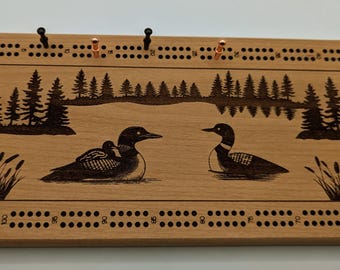 May include: A wooden cribbage board with a nature scene featuring two loons on a lake. The board has a traditional layout with numbered holes and pegs. The scene is engraved into the wood and features a forest background.