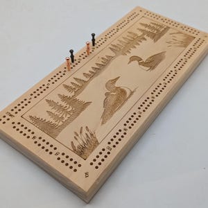 May include: A wooden cribbage board with a nature scene featuring two loons and pine trees. The board has a light wood finish and is decorated with a laser-engraved design. The board has a "START" label and numbered holes for pegs.