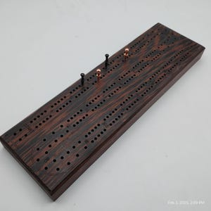 May include: A wooden cribbage board with a dark brown wood grain finish. The board has a track with numbered holes and a start position. There are black and copper pegs on the board.