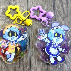 May include: Two acrylic keychains featuring cartoon characters with blue hair and yellow eyes. One character wears a yellow apron and holds a paint palette and brush with a yellow star clasp. The other wears a blue maid outfit and holds a cupcake with a purple star clasp.