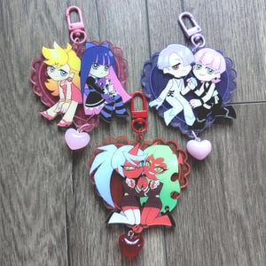 May include: Three heart-shaped acrylic keychains featuring anime-style characters. The top left shows two figures with yellow and blue hair, the top right shows two figures with white and pink hair, and the bottom shows two red-skinned figures.