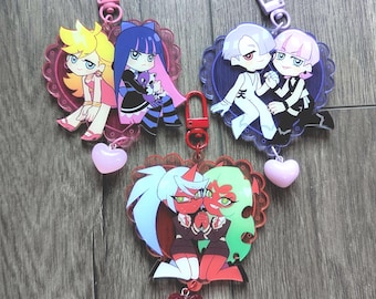 Może przedstawiać: Trzy breloki akrylowe w kształcie serca z postaciami w stylu anime. Po lewej u góry dwie postacie z żółtymi i niebieskimi włosami, po prawej u góry dwie postacie z białymi i różowymi włosami, a na dole dwie postacie z czerwoną skórą.