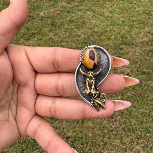 May include: A silver-toned pendant with a large oval-shaped, tan and brown stone at the top. Below the stone is a gold-toned figure with outstretched arms. The pendant has a decorative leaf design at the bottom.