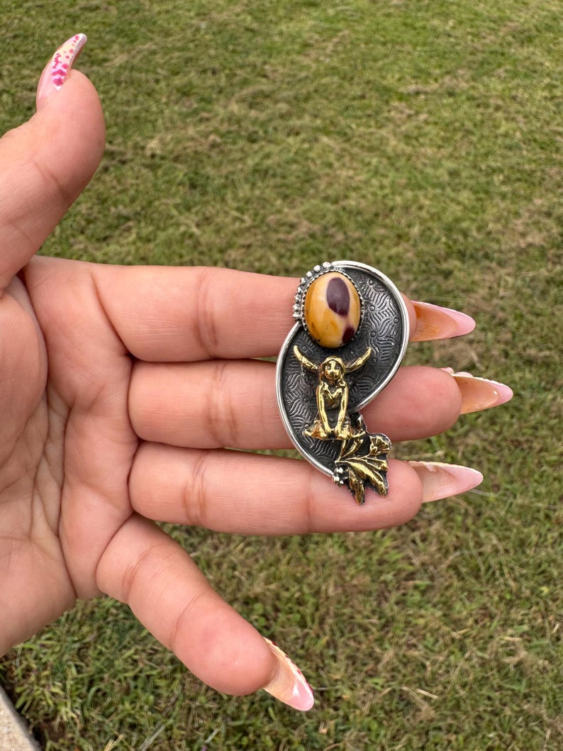 May include: A silver-toned pendant with a large oval-shaped, tan and brown stone at the top. Below the stone is a gold-toned figure with outstretched arms. The pendant has a decorative leaf design at the bottom.