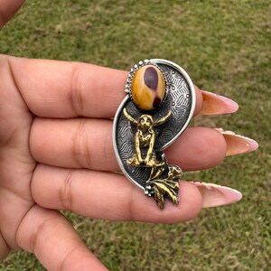 May include: A silver-toned pendant with a central oval-shaped stone in shades of yellow and brown. The pendant features a gold-colored figure and leaf accents. The design is set against a textured background, creating a unique jewelry piece.