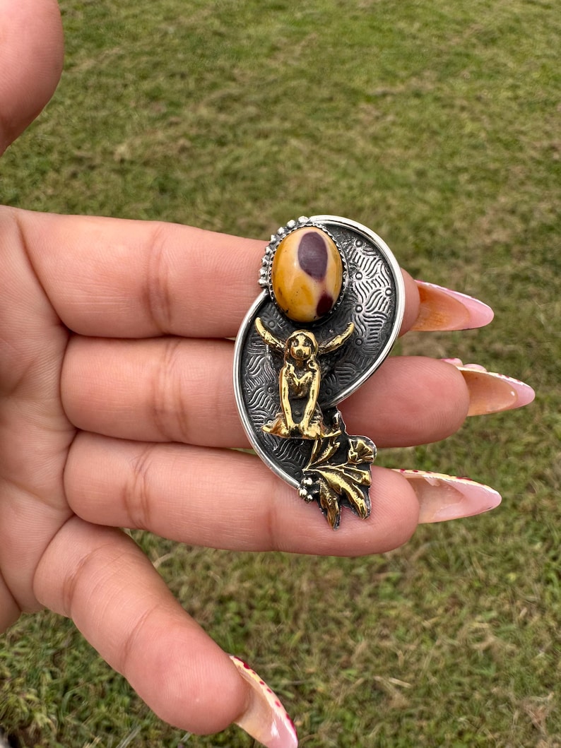 May include: A silver-toned pendant with a central oval-shaped stone in shades of yellow and brown. The pendant features a gold-colored figure and leaf accents. The design is set against a textured background, creating a unique jewelry piece.