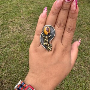 May include: A silver ring with a large, oval, yellow and brown stone. The ring features a small, gold-colored figure and decorative silver accents. A red, blue, and silver bracelet is also visible.