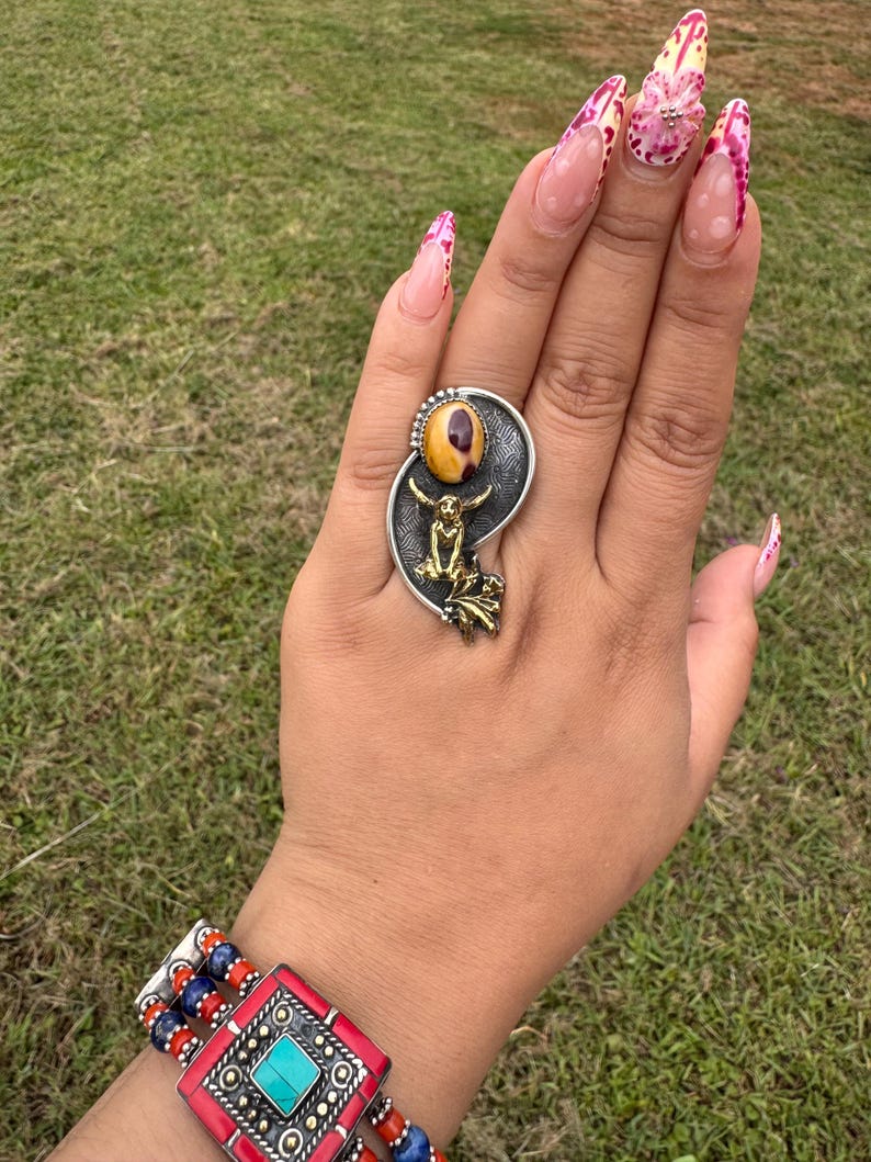 May include: A silver ring with a large, oval, yellow and brown stone. The ring features a small, gold-colored figure and decorative silver accents. A red, blue, and silver bracelet is also visible.