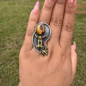 May include: A silver ring with a large oval-shaped, yellow and brown stone. The ring features a gold-colored figure and leaf design. The ring is on a hand with long, decorated fingernails.
