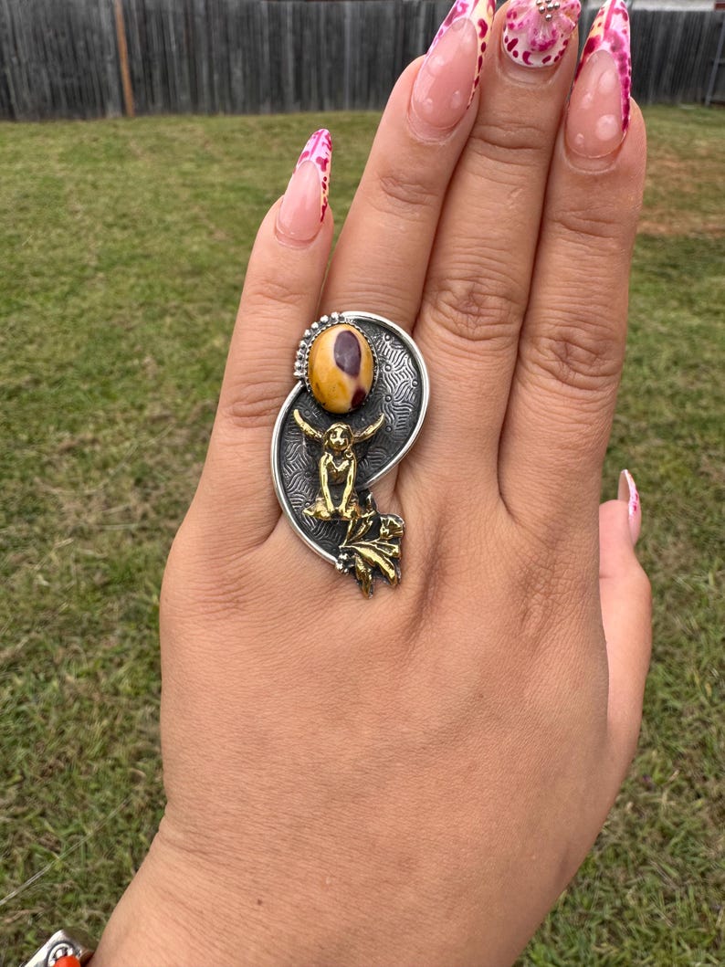 May include: A silver ring with a large oval-shaped, yellow and brown stone. The ring features a gold-colored figure and leaf design. The ring is on a hand with long, decorated fingernails.