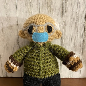May include: A crocheted doll wearing a green sweater, black pants, and a blue face mask. The doll has a tan face, white hair, and black eyes.
