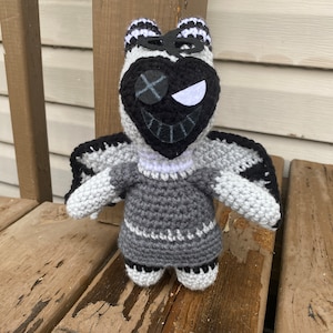 Puede incluir: Un juguete de peluche tejido a crochet con una cabeza negra en forma de corazón, ojos blancos y alas grises. El juguete lleva un vestido gris y blanco.