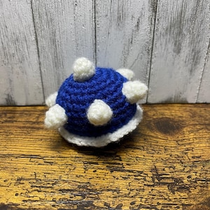 May include: A handmade crochet toy in the shape of a blue and white Koopa shell. The shell has a rounded shape with white, fluffy accents and a white trim. The toy is sitting on a wooden surface.