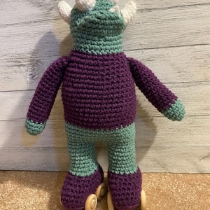 Op de afbeelding: Een gehaakt monster speelgoed van paarse en groene draad, met paarse en groene rolstoeltjes met houten wielen. Het monster heeft witte hoorns en grote witte ogen.