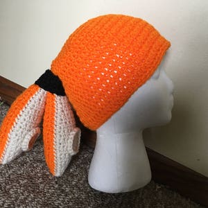 May include: An orange crochet hat with matching appendages, featuring white and black accents. The hat is designed to resemble a character from a video game. The hat is displayed on a white mannequin head.