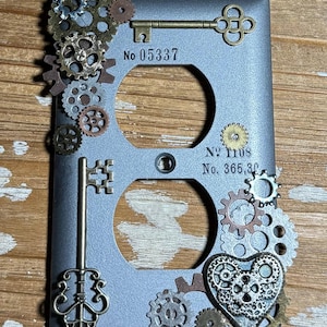 May include: A silver metal outlet cover with a steampunk design. It features gears, keys, and a heart-shaped design. The outlet cover has the numbers "No. 05337", "No. 1108", and "No. 365.30" printed on it.