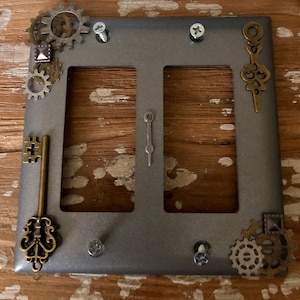May include: A gray metal double light switch cover with decorative steampunk elements, including keys, gears, and clock hands.
