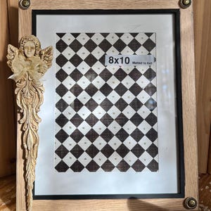 May include: A framed artwork with a black and white checkered pattern, labeled "8x10" and matted to 6x8. The frame is light brown with decorative corner accents. An ornate angel figurine with a gold bow is attached to the frame.