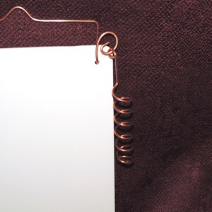 May include: A white paper with a copper wire hanger and a copper coil hanging from the top. The paper is against a dark red background.