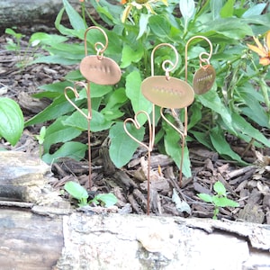 Puede incluir: Tres marcadores de plantas de cobre con acentos de alambre en forma de coraz&oacute;n. Los marcadores est&aacute;n grabados con las palabras "Albahaca", "Romero" y "Tomillo".