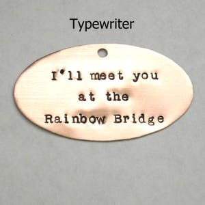 Puede incluir: Una etiqueta ovalada de cobre con el texto "I'll meet you at the Rainbow Bridge" grabado en ella. La etiqueta tiene un agujero en la parte superior para colgarla.