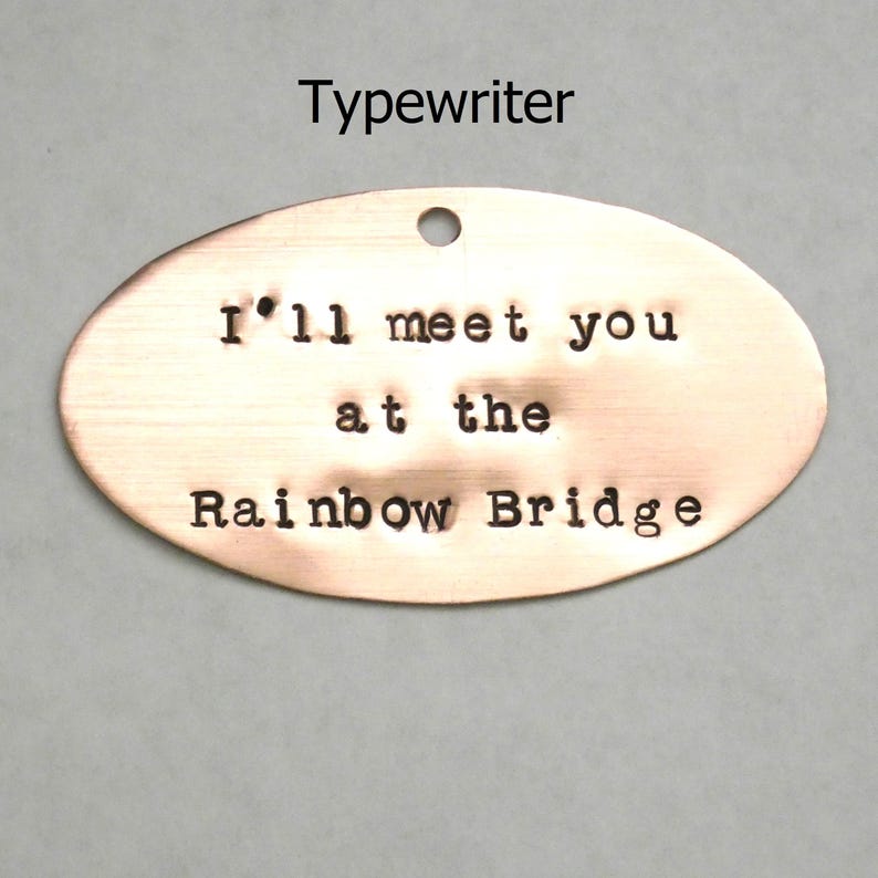 Puede incluir: Una etiqueta ovalada de cobre con el texto "I'll meet you at the Rainbow Bridge" grabado en ella. La etiqueta tiene un agujero en la parte superior para colgarla.