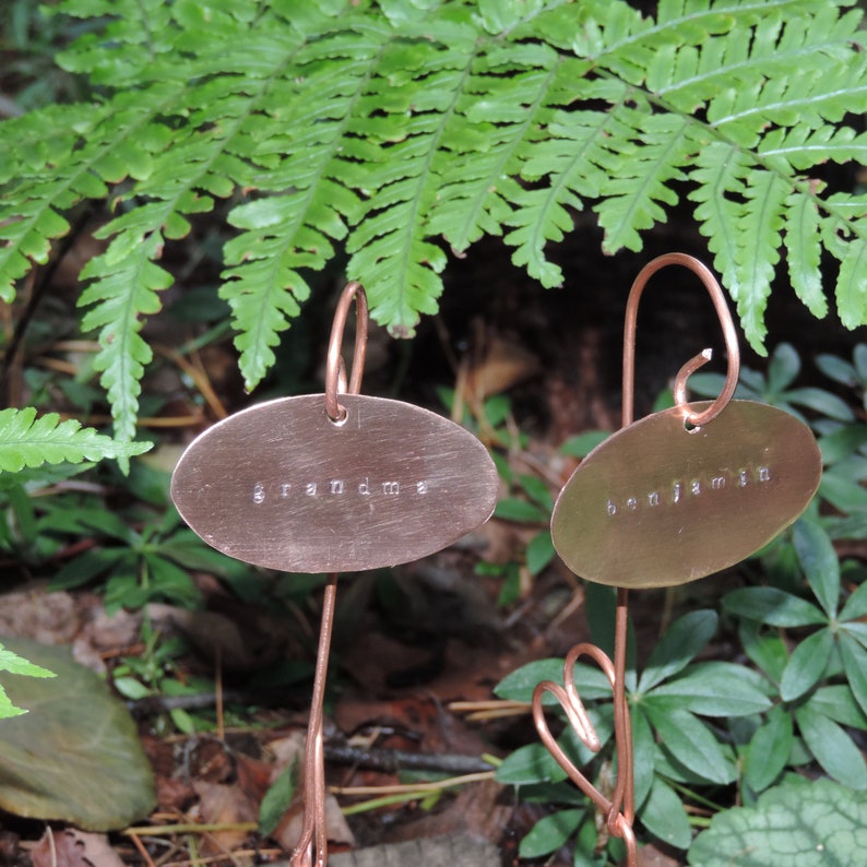 Puede incluir: Dos marcadores de plantas de cobre con etiquetas ovaladas. La etiqueta de la izquierda dice "grandma" y la etiqueta de la derecha dice "benjamin".