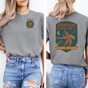 May include: Gray t-shirt featuring a front and back design. The front design includes the text "GUARDIANS OF THE GALAXY" and "COSMIC REWIND" with a starship graphic. The back design features the same text and a larger starship graphic.