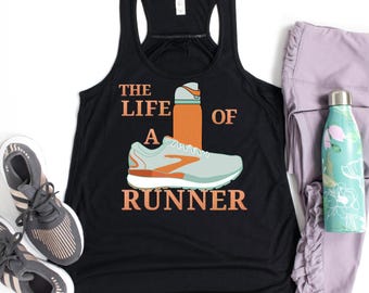 May include: Black racerback tank top with the text "THE LIFE OF A RUNNER" in orange. The design includes a running shoe and a water bottle. Gray running shoes and lavender leggings are also visible.
