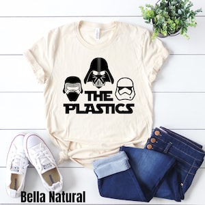May include: A beige t-shirt with a black graphic of three Star Wars characters, Darth Vader, Kylo Ren, and a Stormtrooper, with the text "The Plastics".