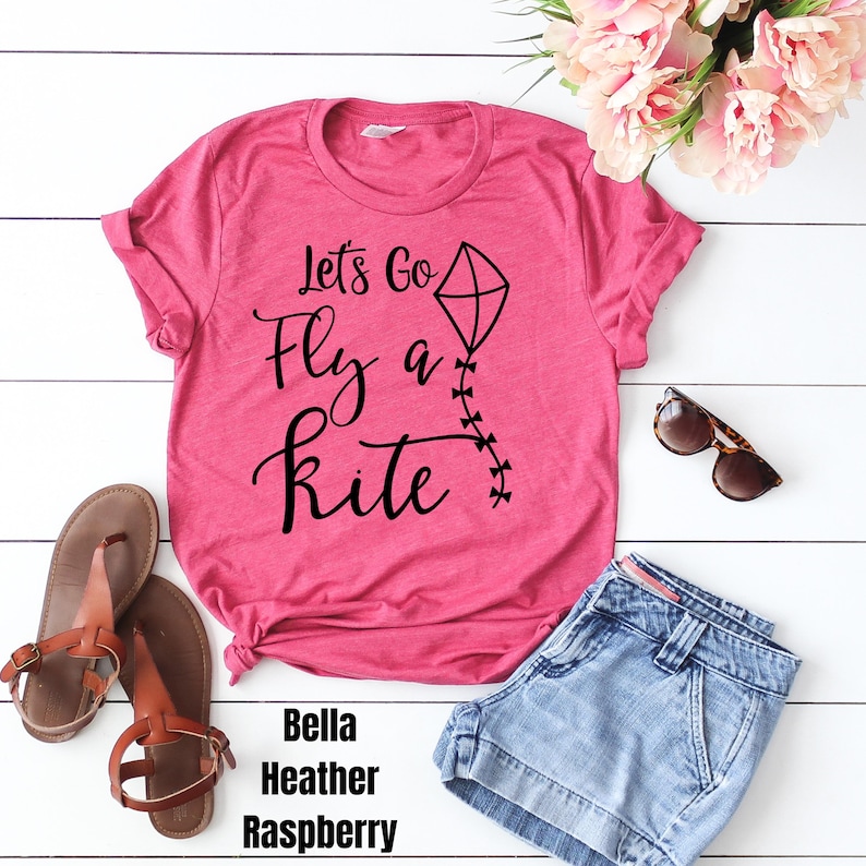 May include: Pink t-shirt with the text "Let's Go Fly a Kite" in black lettering. The shirt features a black kite graphic with a string of black Xs.