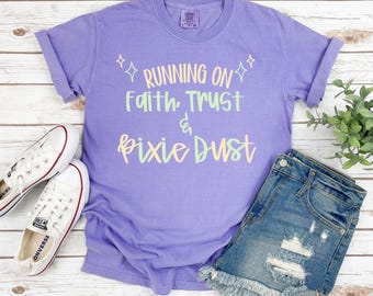 May include: A lilac-colored t-shirt with the text "RUNNING ON Faith, Trust & Pixie Dust" in pastel colors. The shirt is paired with denim shorts and white canvas sneakers. The bottom right corner of the image has the text "CC Violet".