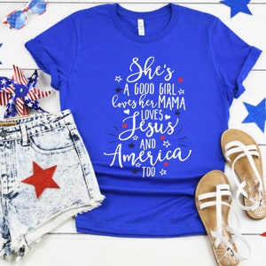May include: A blue t-shirt with a white graphic that says "She's a good girl loves her mama loves Jesus and America too". The shirt is laid out on a white wooden surface with red, white, and blue stars and a denim shorts with a red star.