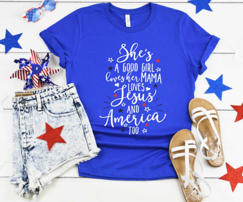 May include: A blue t-shirt with a white graphic that says "She's a good girl loves her mama loves Jesus and America too". The shirt is laid out on a white wooden surface with red, white, and blue stars and a denim shorts with a red star.