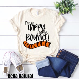 May include: A beige t-shirt with the text "I'm so happy I could bounce!" in black. A black and orange tiger tail graphic is below the text. The shirt is laying on a white surface with a pair of blue jeans and white sneakers.