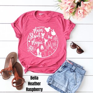May include: Pink t-shirt with a white graphic design that says "Main Street Magic" and features various Disney icons. The shirt is laid out on a white surface with a pair of brown sandals and a pair of blue denim shorts.