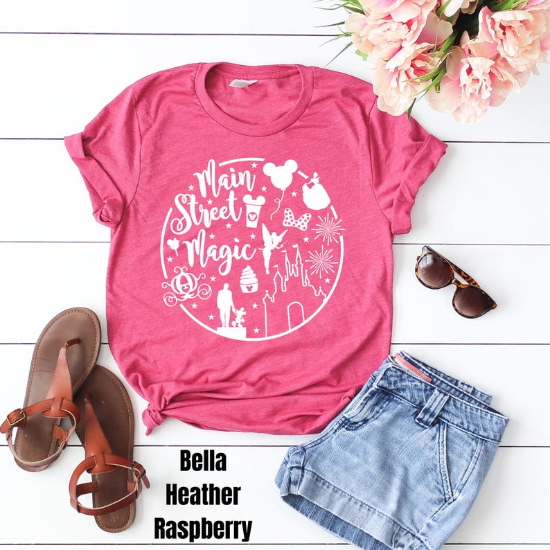 May include: Pink t-shirt with a white graphic design that says "Main Street Magic" and features various Disney icons. The shirt is laid out on a white surface with a pair of brown sandals and a pair of blue denim shorts.