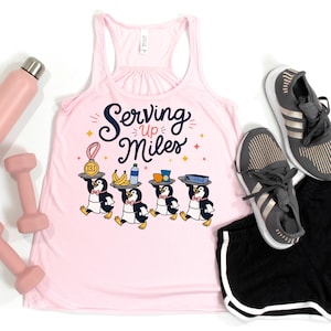 May include: A light pink racerback tank top featuring a graphic of four penguins in bow ties carrying trays with a 13.1 medal, bananas, a water bottle, and snacks, with the text "Serving Up Miles" printed in dark blue script above the penguins.
