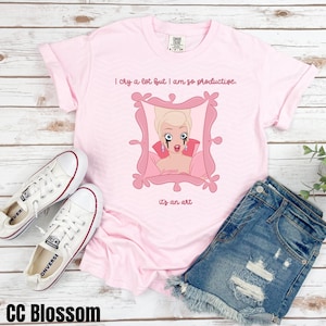 May include: Pink t-shirt with a cartoon image of a woman with blonde hair and blue eyes. The woman is wearing a pink dress and has tears running down her face. The text on the shirt reads "I cry a lot but I am so productive. It's an art."