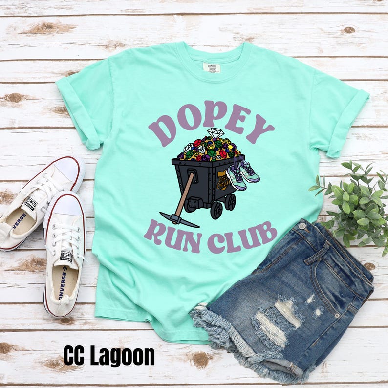May include: A turquoise blue t-shirt with the text "Dopey Run Club" in purple lettering. The design features a black mine cart filled with colorful jewels and a pickaxe.