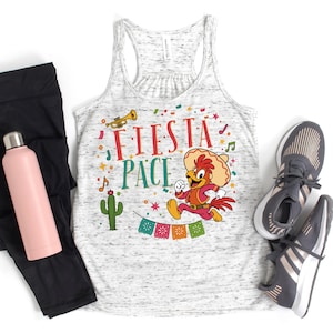 May include: A white heathered racerback tank top featuring a graphic of a cartoon rooster wearing a sombrero, the text FIESTA PACE, a trumpet, a cactus, and colorful bunting. The design includes musical notes and confetti in red, green, and yellow.