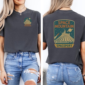 May include: Dark gray t-shirt featuring a Space Mountain graphic on the front and back. The front graphic is a circular design with the words "Space Mountain". The back graphic is a rectangular design with the words "Space Mountain" and "Spaceport".