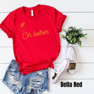 May include: Red t-shirt with a yellow bee and honeycomb design and the text "Oh, bother."  The shirt is paired with denim shorts and white sneakers.