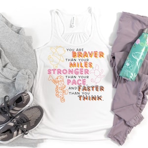 May include: White tank top with the text "YOU ARE BRAVER THAN YOUR MILES, STRONGER THAN YOUR PACE, AND FASTER THAN YOU THINK." The shirt has cartoon illustrations of Winnie the Pooh characters. Gray athletic shoes and leggings are also in the image.