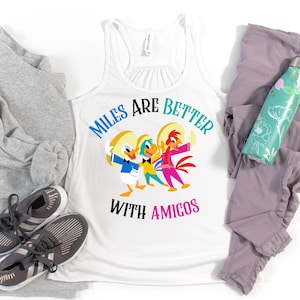 May include: A white racerback tank top featuring a graphic of three cartoon birds wearing sombreros with the text "MILES ARE BETTER WITH AMIGOS" printed in blue, teal, and pink. The design includes Donald Duck, José Carioca, and Panchito Pistoles.
