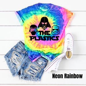 Puede incluir: Una camiseta tie-dye con un esquema de color arco&iacute;ris ne&oacute;n. La camiseta presenta un gr&aacute;fico de tres personajes de Star Wars: Darth Vader, Kylo Ren y un Stormtrooper. El texto "The Plastics" est&aacute; impreso debajo de los personajes.