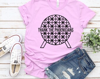 May include: A Bella Lilac t-shirt with the text "THANK THE PHOENICIANS" in black. The graphic is a geometric design. The shirt is paired with denim shorts and white sneakers.